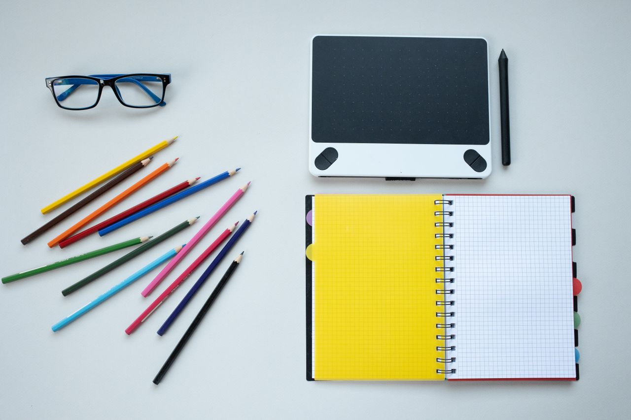 Flat lay of a creative workspace featuring colored pencils, notebook, and graphic tablet.